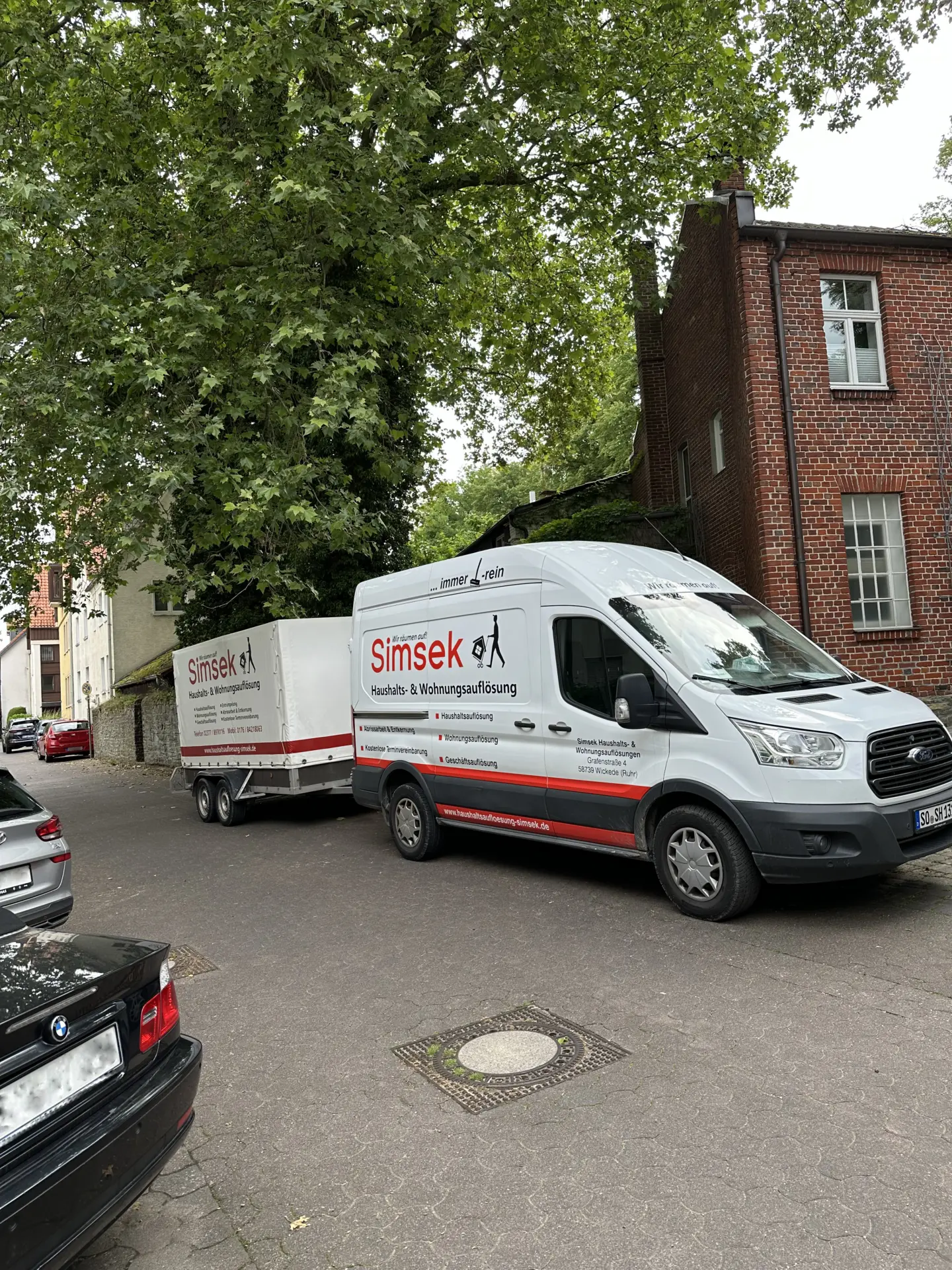 Transporter von Simsek für Haushaltsauflösungen, ausgestattet mit einem Anhänger, auf einer Straße geparkt.