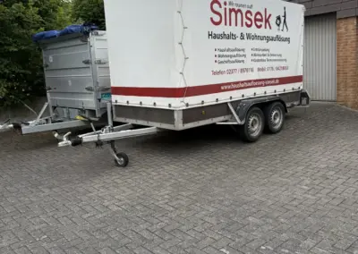 Ein Anhänger mit der Aufschrift 'Simsek' für Haushalts- und Wohnungsauflösungen, geparkt auf einem gepflasterten Platz.